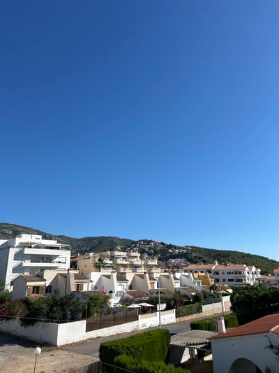 A wide-angle, eye-level shot captures a view of a residential area in Alcalà de Xivert, Spain, nestled at the base of a green, hilly landscape under a clear blue sky. The foreground features a paved area with a small, white lamppost, bordered by a low white wall and dense green hedges. Adjacent to this is a road and more hedges.

Dominating the mid-ground is a collection of white, multi-story buildings with terracotta tile roofs. These structures are characterized by their angular designs, white balconies, and occasional awnings. Some buildings are tightly clustered, while others stand slightly apart, creating a tiered appearance that follows the gentle slope of the land. Vegetation, including trees and flowering bushes, is interspersed among the buildings, with some plants reaching up to balcony level. A tall palm tree stands out near the center-right of the building cluster.

The background is defined by the undulating hills covered in lush green vegetation. Scattered across the hillsides are more buildings, appearing smaller from this distance, suggesting a sprawling residential development climbing the slopes. The overall scene is bathed in bright sunlight, casting minimal shadows and indicating a clear day. No people are visible in the frame.
