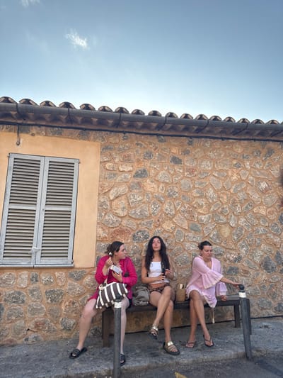 Three young adult females are seated on a dark-colored wooden bench against a building in Deià, Spain. The building features a multi-toned stone wall composed of irregular brown, grey, and reddish stones. To the left, a section of the wall is covered with smooth, light orange stucco, containing a closed window with light grey louvered shutters. A black electrical cable runs horizontally across the stone wall below the roofline. Above the wall, a dark corrugated gutter and a row of terracotta roof tiles are visible under a clear, light blue sky with a few wispy white clouds.

From left to right on the bench: The first female wears a fuchsia-colored long-sleeved top or dress and black sandals. She holds a small white rectangular object in her hands and has a white flower in her hair above her right ear. A brown and white striped tote bag rests next to her on the bench. The middle female wears a white spaghetti-strap top and patterned shorts, with long dark hair. She holds a black smartphone in her right hand and wears gold-colored sandals. Her legs are crossed. The third female wears a light pink loose-fitting dress with wide sleeves and dark flip-flops. Her right hand is extended towards the end of the bench, and a small woven bag is placed next to her. The ground in the foreground is dark paving with a faint yellow line partially visible beneath the bench.