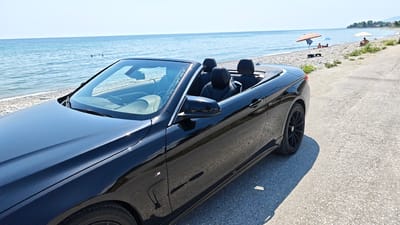 A black convertible BMW 4 Series is parked on a paved surface next to a pebble beach. The car's top is down, revealing black leather seats. The ocean stretches out in the background, with calm blue water and a clear sky. Two people are visible swimming in the sea. Further down the beach, several individuals are relaxing under beach umbrellas, suggesting a sunny day. The scene is likely set in Leptokarya, Greece, given the location context. The time of day appears to be daytime, possibly late morning or early afternoon, judging by the bright sunlight and clear visibility. The overall mood is relaxed and summery. There is a small "M" badge on the side of the car, indicating it is an M Sport model.