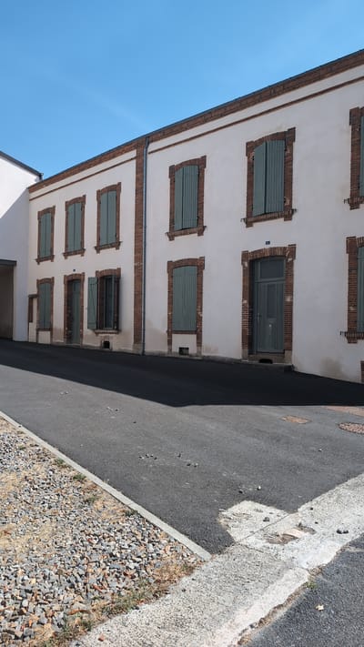 The image shows a close-up view of a building's facade and the adjacent street in the city of Cholet, France. The building has a white plaster exterior with red brick accents around the windows and at the roofline. Several windows with green shutters are visible, as well as a grey double door. The street in front of the building is paved with asphalt and bordered by a gravel area. The sky is a clear, bright blue. The scene appears to be a sunny day.