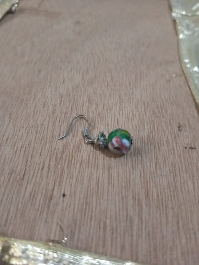 A single earring is depicted lying on a light brown wooden surface with visible vertical grain lines. The earring consists of a silver-colored wire hook component attached to a spherical bead. The bead is approximately 8-10mm in diameter, primarily green, featuring red and white floral or abstract patterns. The silver-colored components show minor signs of wear or tarnish. The earring is positioned slightly off-center to the right. Out-of-focus, reflective, possibly metallic or fabric elements are visible along the top and bottom edges of the frame. The scene is located in Chandragiri, Nepal.