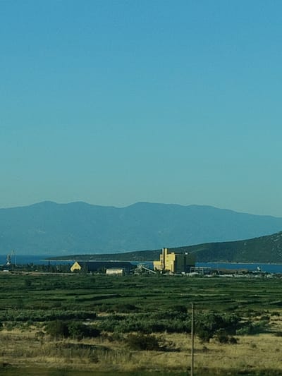 The image captures a wide view of a coastal industrial area. In the foreground, a dry, grassy field with some scattered bushes stretches towards the middle ground. The middle ground reveals a lush, green agricultural landscape with terraced fields and rows of vegetation. Dominating the middle ground and extending into the background is a body of water, likely the sea, with a distant coastline featuring rolling, green hills.
On the coastline, several industrial structures are visible. A prominent yellow building, possibly a factory or processing plant, stands out. Adjacent to it are other industrial buildings, some appearing to be warehouses or storage facilities, and what looks like a dock or loading area. A tall crane is also visible to the left of the yellow building. The background is characterized by a range of soft blue mountains under a clear, light blue sky.
The scene appears to be located in the Municipal Unit of Almyros, Greece, given the geographical characteristics of the area. It is a daytime scene, with clear lighting and no visible human presence or activity. The overall impression is one of a functional industrial zone integrated into a natural coastal landscape.