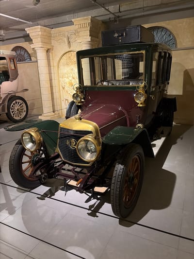 An antique automobile, likely from the early 20th century, is the central subject of this indoor scene, possibly within a museum or grand hall in Wassenaar, Netherlands. The car features a two-tone paint scheme with a deep maroon body and dark green fenders and trim. Its front is adorned with gold-colored brass accents, including two large, prominent circular headlights and two smaller side lamps. The radiator grille, also brass-colored, showcases a distinct dark oval emblem in its center, displaying what appears to be a stylized "SP" monogram.
The car is equipped with wooden-spoked wheels fitted with black tires, and a spare tire is mounted prominently on its right side. A black, suitcase-like trunk is attached to the rear of the vehicle. In the background, ornate architectural elements are visible, including beige stone columns, arches, and decorative relief carvings depicting foliage and possibly figures. A decorative metal grille is also seen behind the car's rear.
A partial view of a second, lighter-colored vintage car with white spoked wheels is visible in the lower-left background, suggesting a collection or exhibition of historical vehicles. Strong artificial lighting casts distinct shadows of the main car and its components onto the light-colored floor. The scene depicts a static display, highlighting the craftsmanship and design of vintage automobiles within an architecturally rich and historic environment.