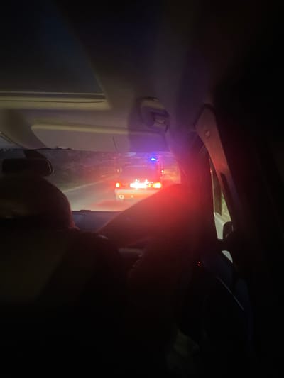 The image is a night-time shot from inside a vehicle, looking forward through the windshield. The interior of the vehicle is dark, with a sunroof visible at the top and parts of the dashboard and pillars. In the distance, an emergency vehicle, possibly an ambulance or police car, is stopped on a road. It has flashing red and blue lights. The road appears to be in a rural or semi-rural setting, not clearly within a city. The presence of flashing lights suggests an ongoing incident or a hazard on the road. There is no specific visual information to confirm the location is Beloci, Moldova.