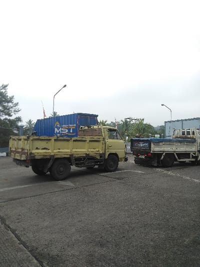 The scene depicts an outdoor industrial or commercial area in Cijedil, Indonesia, under an overcast sky. A weathered yellow two-axle light-duty truck is positioned on the left, angled towards the right, appearing to be in transit or recently halted. Its flatbed is loaded with a blue shipping container, approximately 20 feet in length, marked with the white stencil "MST" on its visible side. To the right, a second light-colored, two-axle light-duty truck with a blue tarp covering its bed is stationary, its rear facing the viewer. Both vehicles rest on a grey, worn asphalt or concrete pavement exhibiting faded white line markings. The background features scattered green trees, including palm trees, two dark-colored street light poles, and indistinct industrial or commercial structures. No individuals are visible in the frame.