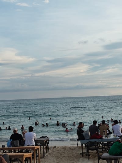 The image depicts a beach scene in Monikeun, Indonesia. In the foreground, several groups of people sit at tables on the sand, seemingly at a beachside establishment. Behind them, numerous people are swimming and playing in the ocean. The sky is overcast with a mix of light blue and grey clouds. The overall setting suggests a relaxed, leisure-oriented atmosphere.