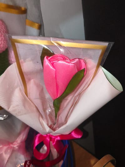 A close-up shot captures a single, bright pink artificial rosebud, its petals made of shimmering satin. The rose is encased in clear plastic wrapping, edged with a thin gold ribbon, and presented in a light pink paper wrapper. A green satin leaf juts out from the base of the rose. The bouquet is tied with a vibrant pink satin ribbon. In the background, out-of-focus elements suggest other similar rose bouquets, some with pink pompoms. The lighting is dim, creating a sense of intimacy. The overall impression is one of a handcrafted gift. There is no visible text in the image. The context provided suggests this image was taken in Cavite City, Philippines.