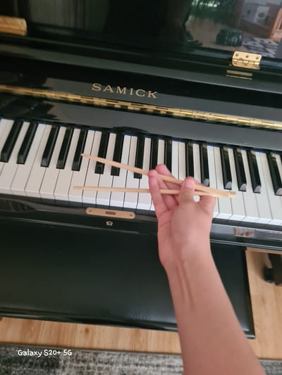 An overhead shot shows a person's right arm and hand holding two wooden chopsticks over the black and white keys of a black upright piano. The "SAMICK" logo is visible on the piano's casing. The piano is situated in an indoor setting. A watermark reading "Galaxy S20+ 5G" is present in the bottom left corner of the image. There is no information to suggest the image was taken in Woodburn, United States of America.