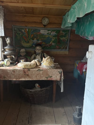 The image depicts the interior of a rustic dwelling, likely a izba, in the village of Bol'shoi Mashlyak, Russia. The walls and ceiling are constructed from rough-hewn logs. A table covered with a floral tablecloth is centrally positioned, laden with various items. On the table, a large round loaf of bread, wrapped in plastic, is prominent. To its left, a vintage alarm clock displays approximately 10:15. A samovar, a traditional Russian metal container for heating and boiling water, stands on a surface to the left of the table. Two mannequin figures, appearing to be elderly individuals dressed in traditional attire, are seated behind the table. The male figure wears a cap and a vest over a white shirt, while the female figure is adorned with a headscarf and a colorful dress. A teacup with a saucer is placed in front of the female figure. On the right side of the table, a mound of what appears to be popcorn or a similar snack is displayed in plastic packaging. Beneath the table, a woven basket contains a white duck with an orange beak, seemingly nestled in straw. To the right of the table, a bed with colorful bedding is visible. A tapestry depicting a pastoral scene with figures and animals hangs on the log wall above the table. A clock with a round face is mounted on the wall above and to the left of the tapestry. The floor is made of wooden planks. A dark, metal container with a handle, possibly an old oil lamp or jug, sits on the floor in the foreground to the right. Green and white curtains are draped near the ceiling on the right side of the room. The overall scene suggests a preserved or staged representation of traditional rural life.