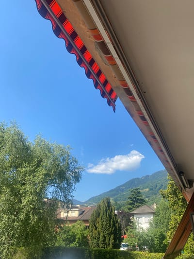 The image depicts a view upwards from under a striped awning towards a blue sky with a single white cloud. In the distance, mountains covered in trees are visible. Buildings are interspersed among the greenery. The scene is likely located in Monthey, Switzerland, given the presence of Alpine scenery and architecture typical of the region. There are no people depicted.