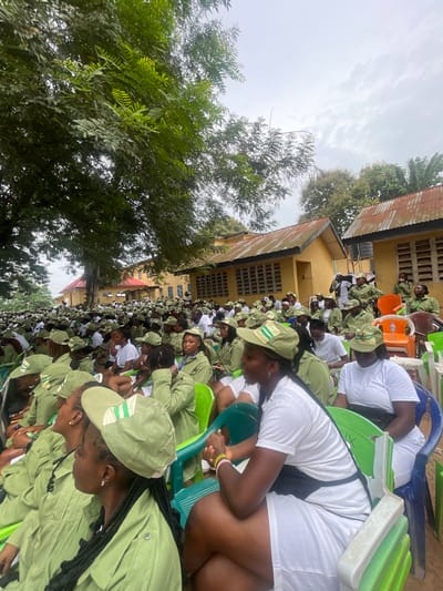 This photograph, taken outdoors, captures a large group of young people, predominantly women, seated on green and blue plastic chairs. They are attending an event, likely a briefing or assembly, given their uniform attire. The majority are dressed in light green shirts and trousers, with matching caps adorned with green and white stripes. Some individuals wear white t-shirts and shorts. The setting appears to be a communal area, with yellow buildings featuring corrugated iron roofs in the background. A large tree with dense foliage provides shade over a portion of the gathering. The overall atmosphere suggests an organized event within a Nigerian context, possibly related to a national youth service or educational program. The location is Emure-Ekiti, Nigeria, a town in Ekiti State known for its participation in such programs.