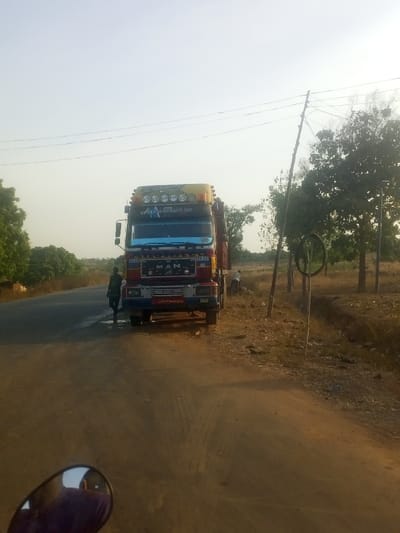 This image depicts a large red and blue truck, likely a tipper or dump truck, stopped on the side of a road in what appears to be a rural or semi-urban area. The truck is parked partially on a dirt shoulder and partially on a paved section of the road.
There are at least two people visible. One person, dressed in dark attire, is standing near the front driver's side of the truck, seemingly looking at or interacting with the vehicle. Another person is further away on the right side of the image, near the truck's rear, stooping down in the grassy area alongside the road.
The setting is characterized by a dirt road in the foreground leading into a paved road where the truck is situated. Sparse trees and dry vegetation line the sides of the road, particularly prominent on the right. A wooden utility pole with power lines stretches across the upper part of the frame, and a distinct black, circular object, possibly a convex traffic mirror or part of some equipment, is visible on a post on the right. A motorcycle's rearview mirror is partially visible in the bottom left corner, suggesting the photo was taken from a motorcycle.
The time of day appears to be late afternoon or early evening, given the warm, soft light and the clear, bright sky. The weather is sunny with no clouds.
Visible text on the truck includes the brand name "MAN" on its front grille. Above the windshield, a website address "www.bm&sons.com" is displayed. Below the grille, the phrase "Life's Changes" is written. The scene portrays a casual stop or an activity related to the truck on a quiet road in Nunku, Nigeria.