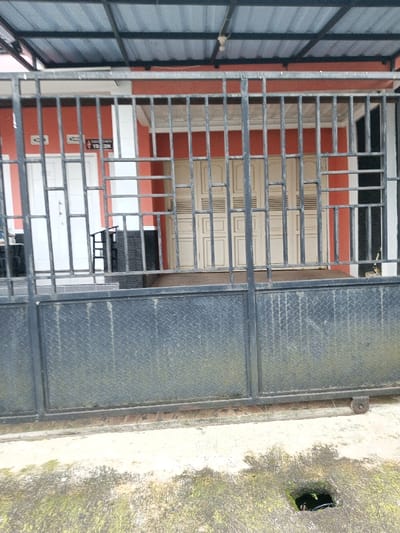 The image depicts the exterior of a building in Viengthong, Laos, viewed from behind a dark grey metal sliding gate.

In the foreground, a concrete ground surface shows patches of green moss and a square drain opening with visible darkness inside. The sliding gate spans the width of the frame, consisting of a lower half constructed from dark grey diamond plate metal and an upper half featuring a grid of vertical and horizontal bars. The gate's wheels are visible at its base on the right.

Behind the gate, a building with an orange-red facade is visible under a dark grey corrugated metal roof supported by a visible framework. On the left side of the building, a white single door is present. Above this door, two white rectangular signs depict restroom symbols: a male figure on the left and a female figure on the right. Below the female figure, partially visible text appears to read "TOILET" with "YOLON" underneath. To the right of the white door, a dark, possibly black, object resembling a stack of chairs or a piece of furniture is situated. Further to the right, a set of light-colored, possibly cream or beige, double doors with a multi-panel design serve as another entrance. A light fixture hangs from the underside of the roof structure above the doors. No individuals are present in the scene.