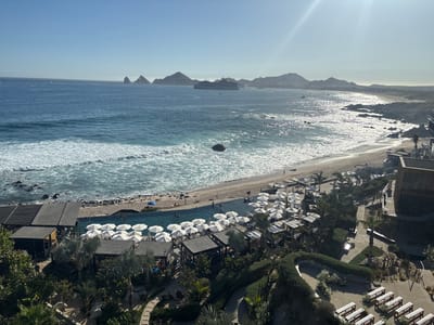 This is an outdoor scene at a coastal resort in Cabo San Lucas, Mexico on a sunny day. The foreground shows manicured gardens with palm trees, hedges, and walkways. An area is set up with benches and floral arrangements, possibly for a wedding or event. A swimming pool with white umbrellas and lounge chairs borders the beach. People are swimming in the pool and walking on the beach. Beyond the beach is the ocean with waves breaking. In the distance, mountains can be seen along the coastline. A large cruise ship is anchored offshore. The sky is clear with a bright sun.