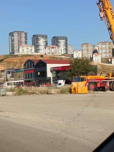 In Yahşihan, Turkey, a construction site unfolds under a clear blue sky. A large yellow Liebherr crane dominates the right side of the frame, its arm extending upwards and towards the left. In the foreground, a yellow and red crane truck is parked, bearing the phone number "(0542) 807 04 72" and the "LIEBHERR" logo. Several red tractors are lined up in front of a modern building with a red roof. Behind these vehicles, a multi-story building with dark windows and a red section is visible. Further in the background, a series of tall residential buildings with white and gray facades rise against the skyline, situated on a dirt-covered hill. The ground is mostly unpaved, with patches of dry grass and scattered stones. A metal dumpster is present on the far left of the image. The scene captures a moment of ongoing construction and development within the city.
