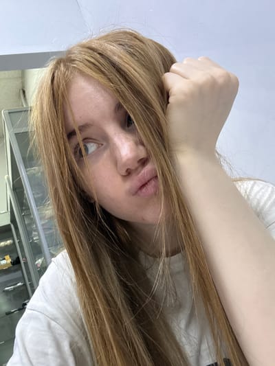 A young woman with long, light brown hair is captured in a close-up, selfie-style photograph. Her left hand is propped on her forehead, and her face is turned slightly to her left, with her lips puckered in a pout. She is wearing a white t-shirt. In the background, a glass-fronted cabinet with shelves is visible, containing various items that appear to be displayed. The setting suggests an indoor location, possibly a shop or an exhibition space, with a plain, light-colored wall behind the cabinet. There is no specific indication of the city of Kuznetsk, Russia within the image itself.