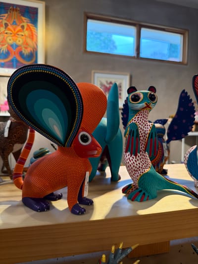 A collection of brightly colored, wooden animal carvings are displayed on a light wooden surface. The foremost carving is an orange creature with a large, teal and blue teardrop-shaped appendage. Above it is a teal carving and another bird-like carving with red and white patterned wings. In the background are a window and framed artworks on a gray wall, adding depth to the scene.