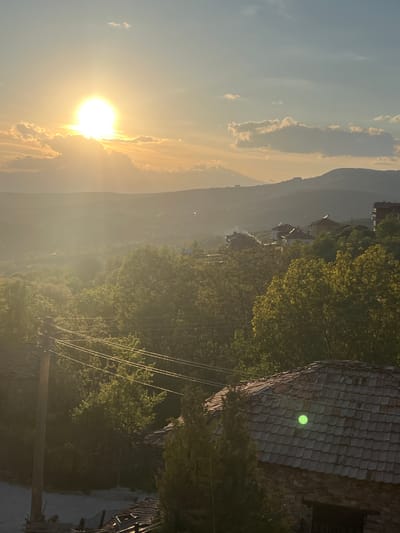 The image depicts a landscape at sunrise or sunset, likely in Kribul, Bulgaria, based on the presence of mountainous terrain and village architecture. The sun is prominently positioned on the left side of the frame, partially obscured by clouds, casting a bright, warm light across the scene.

A mountain range is visible in the background, partially obscured by haze or mist. Below the mountains, a village or town is discernible, with buildings clustered together. A wooden roof is in the foreground on the right side of the scene. Power lines run through the midground, and the presence of trees suggests a partly forested area. The overall impression is of a rural or semi-rural environment.