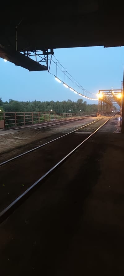 The image depicts an outdoor industrial area with train tracks under a twilight sky. The foreground shows two parallel train tracks running into the distance. A dark, solid structure, possibly a bridge or overpass, partially obscures the top of the image. Suspended from this structure, a series of lights illuminate a pathway or walkway that extends over the tracks. Beyond the tracks, a brown metal fence runs along the perimeter, separating the train area from a dense line of green trees in the background. Some additional lights are visible in the distance, illuminating a large industrial structure, possibly a crane or conveyor system. The overall scene suggests a working industrial or logistical facility. This location is identified as the Salzgitter steelworks in Salzgitter, Germany. There are no people or animals visible in the image.