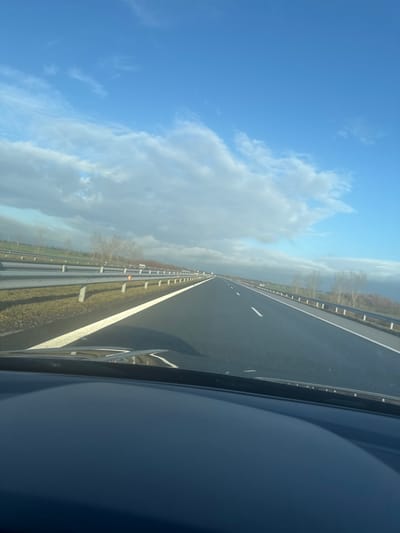 The image displays a forward-facing view of a multi-lane asphalt highway, observed from the interior of a moving vehicle, located near Воденичане, Bulgaria. The road surface, appearing dark and reflective in sections, extends linearly towards the horizon under a sky characterized by a mix of blue patches and scattered white to grey cumulus clouds. The lower portion of the image shows the dark dashboard and the base of the windshield of the photographing vehicle. The highway's rightmost lane is delineated by a continuous solid white line, while dashed white lines mark additional lanes further down the road. Parallel to the highway, on both sides, run metallic guardrails; the right-side guardrail is more prominent and features multiple orange rectangular reflectors. Beyond the guardrails, the landscape comprises grassy verges and fields, punctuated by clusters of bare deciduous trees. No human figures or other active subjects are discernible within the frame.