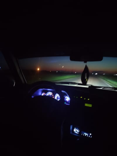 The image shows the interior of a car at dusk or dawn, viewed from the driver's perspective. The car is on a road, possibly a highway, with a dark sky and a strip of light on the horizon. There are lights visible in the distance, suggesting a settlement or road infrastructure. The dashboard is illuminated with blue and red lights from the gauges and a yellow display screen. A car air freshener hangs from the rearview mirror. The scene is dark, with limited visibility of the surroundings outside the car. The location is identified as "Дорожное, Kazakhstan."