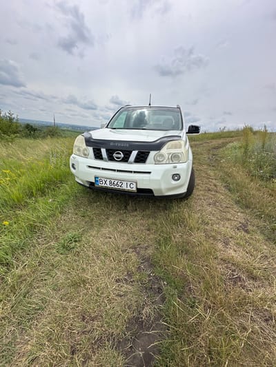 A white Nissan X-Trail is parked on a dirt path in a grassy, hilly area under a cloudy sky. The car's front license plate, reading "BX 8662 IC", is visible. The car has a black bug deflector on its hood. The surrounding environment is overgrown with tall grass and scattered wildflowers, with trees and distant buildings visible in the background. The overall scene suggests a rural or semi-rural setting.
