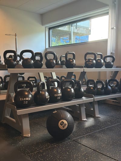 A well-lit gym interior showcases rows of black kettlebells neatly arranged on two tiered metal racks. The kettlebells vary in size and weight, with some displaying "ELEIKO" branding and numerical weight indicators like "16," "20," "32," and "8 kg." In the foreground, a black medicine ball with a gold circular emblem is positioned on the textured, speckled black rubber gym floor. The background features a large window with a view of a modern building and greenery. The overall environment suggests a clean and organized fitness facility.