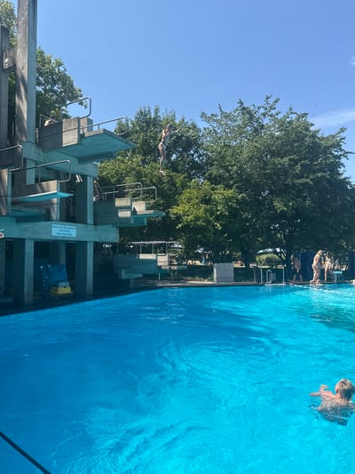 In Rheinbach, Germany, on a sunny day, a young woman is captured mid-air as she leaps from a high diving board at a public swimming pool. The diving structure, made of concrete and metal, stands tall against a clear blue sky, surrounded by lush green trees. Below, the pool's vibrant turquoise water reflects the bright sunlight. In the foreground, another person is swimming in the pool. In the background, other swimmers and sunbathers are visible near the pool's edge. A sign on the diving structure reads "Nicht in den Sprungbereich schwimmen WasserTiefe 3.80m," which translates to "Do not swim in the diving area. Water depth 3.80m."