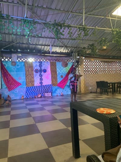 A woman stands against a colorful backdrop with traditional patterns and straw hats. A fan is visible near the ceiling. The background is outdoors with a metal roof structure partially covered with vines. To the right, a dark, patterned table sits on a gray and white checkerboard floor. Black chairs are visible in the background near a patterned brick wall.
