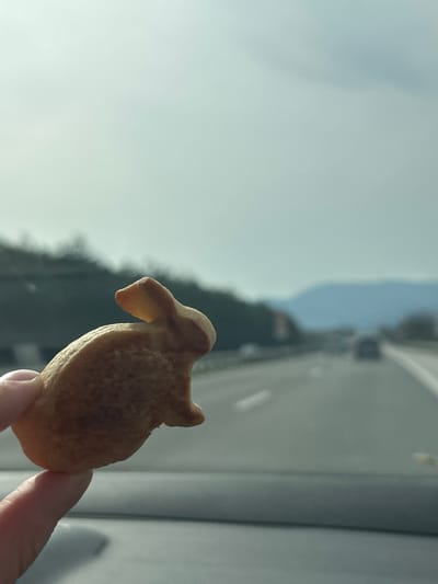 A hand holds a small, tan-colored cookie shaped like an animal, possibly a sheep or a dog, against a blurred background of what appears to be a road, trees, and a cloudy sky. The location in the city of Церово, Bulgaria cannot be determined from the provided image.