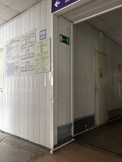 The image depicts a hallway within a building, likely an educational institution given the signage. On the left, a large floor plan titled "ПЛАН БЕЛОВСКОГО ПЕДАГОГИЧЕСКОГО КОЛЛЕДЖА" (PLAN OF BELOVO PEDAGOGICAL COLLEGE) is mounted on the wall. This plan details the layout of the college across three floors, with room numbers and names. Adjacent to this is a smaller sign indicating an emergency exit with a green arrow. Further down the hallway, a door is visible with "ТУАЛЕТ" (TOILET) and "WC" signage. The walls are paneled with white material, and the ceiling appears to be made of acoustic tiles. The floor is a cracked concrete surface with some darker patches. Pipes are visible running along the base of the wall. The overall impression is of an interior hallway in a functional but somewhat dated building. There are no people visible in the image. The presence of the "Belovo Pedagogical College" plan strongly suggests the location is within the city of Belovo, Russia.
