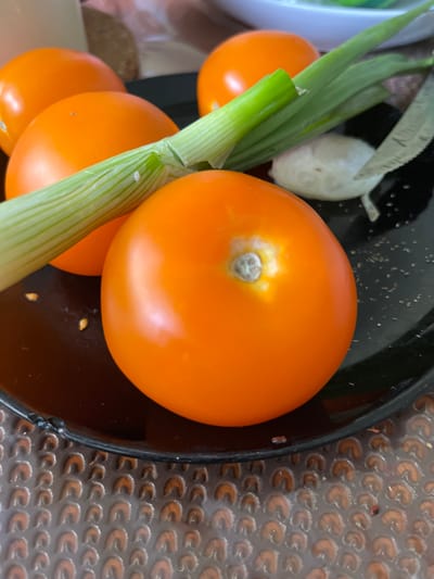 The image shows a close-up view of several bright orange tomatoes, a stalk of green onions, and a piece of garlic on a black plate. The plate is resting on a brown textured surface. The lighting suggests it may be indoors. The tomatoes are round and appear ripe. One tomato in the foreground is particularly prominent, showing its stem end with a slight discoloration. The green onion stalk is positioned across the tomatoes, with its green leaves and white base visible. The garlic piece is small and white, located to the right of the green onions. The overall scene is a simple, still-life arrangement of fresh produce, likely prepared for cooking or serving. There are no people visible in the image, and no specific details indicate the location in Kokkola, Finland.