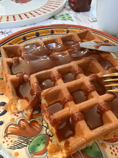 Two waffles, generously coated with caramel sauce, rest on a decorative plate featuring colorful fruit and other motifs. A knife and fork are positioned on the waffles. In the periphery, another plate with a dark chocolate item and holiday-themed napkin are visible. The image presents a close-up, angled view of the food arrangement.