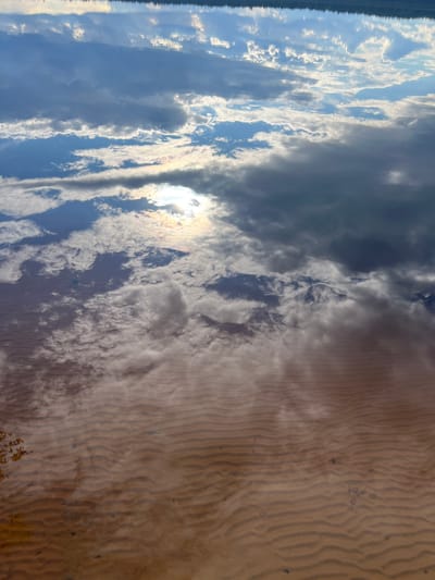 The image shows a reflection of the sky and clouds on the surface of a shallow body of water. The water is clear, allowing the sandy bottom to be visible, with ripples creating a wave-like pattern. The sky reflection is dominated by various cloud formations, some dense and dark, others wispy and white, with patches of sunlight breaking through. The overall impression is one of a tranquil natural scene, likely a lake or a slow-moving river. The location of Дражнаўскі сельскі Савет, Belarus is provided, suggesting this is a scene from that region.
