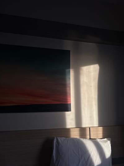The image depicts a bedroom scene with a focus on a wall adorned with a landscape artwork and illuminated by dramatic natural light casting shadows. The artwork, a rectangular canvas, displays a gradient of colors transitioning from deep teal at the top to warm reds and oranges, below which is a dark, silhouetted landscape of hills. To the right of the artwork, a wall is illuminated by shafts of light, creating distinct rectangular and elongated shapes of brightness against a darker background. The lighting suggests a low sun angle, likely from a window. Below the artwork and the illuminated wall, the top portion of a wooden headboard is visible, with a white pillow resting on it. The pillow is partially lit by the same natural light, showing soft folds and shadows. A thin, dark vertical object, possibly a book or a remote, is subtly placed behind the pillow. The room appears to be a bedroom, possibly in a hotel or residence, in Surat Thani City Municipality, Thailand. The overall mood is serene and atmospheric due to the interplay of light and shadow.