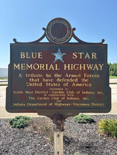 A close-up, eye-level outdoor, daytime photograph shows a dark, rectangular memorial sign mounted on a pole. The sign features raised gold lettering and a decorative crest at the top. The crest has the words "NATIONAL COUNCIL OF STATE GARDEN CLUBS" in a circular arrangement around a relief image of what appears to be a deer. Below the crest, the words "BLUE STAR MEMORIAL HIGHWAY" are prominently displayed in large gold letters. Underneath this, smaller gold text reads: "A tribute to the Armed Forces that have defended the United States of America." Further down, it states, "SPONSORED BY South West District - GARDEN CLUB of Indiana, Inc., IN COOPERATION WITH The Garden Club of Indiana, Inc., AND Indiana Department of Highways - Vincennes District." A small, stylized blue star is positioned above the word "MEMORIAL." The sign is weathered, with some reddish-brown discoloration and faint camouflage patterns visible on its surface. The pole supporting the sign is metal and appears somewhat rusted and worn. In the foreground, the ground is covered with dark wood chips and mulch, with several low-lying green bushes and some dry, brown vegetation. The background shows a bright blue sky and a distant line of green trees. The image appears to be taken in Dale, Indiana, United States, given the text on the sign.