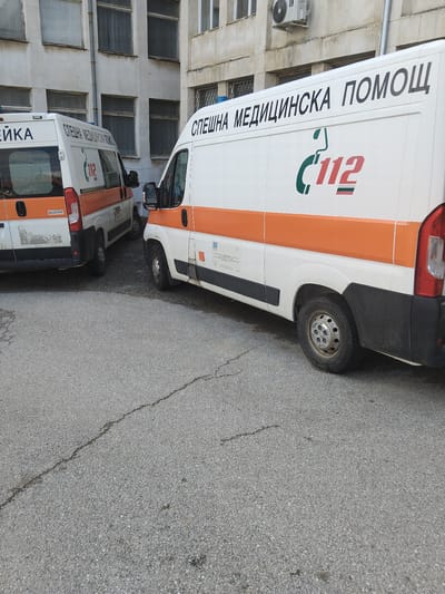 The image depicts two parked white and orange Bulgarian Emergency Medical Aid (BEMS) ambulances in Pirdop, Bulgaria, positioned on an asphalt surface in front of a light-colored institutional building. The ambulance in the foreground, on the right side, is partially visible, displaying "СПЕШНА МЕДИЦИНСКА ПОМОЩ" (Emergency Medical Aid) in black Cyrillic text and a "C112" logo with a green phone receiver and red numerals, underscored by green and red stripes. A second, similar ambulance, identified as a Peugeot Boxer by a rear emblem, is parked behind the first to the left, showing its rear and side with matching livery and text. Blue emergency lights are visible on its roof. The background building is multi-story with numerous rectangular windows, some with blinds, and exhibits weathered facades with an external air conditioning unit. The asphalt ground features visible cracks. No individuals are present in the scene.