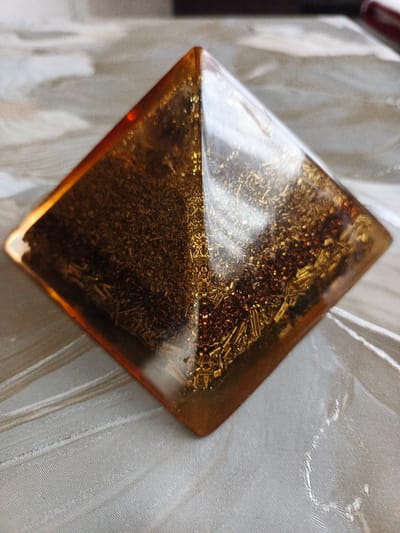 A close-up shot shows an amber-colored, pyramid-shaped object containing metallic particles. The pyramid is sitting on a surface with a subtle, textured pattern. The metallic particles within the pyramid appear to be a mix of golden and brown colors. The photograph was taken in Stroevo, Bulgaria. No people or actions are visible in the image.