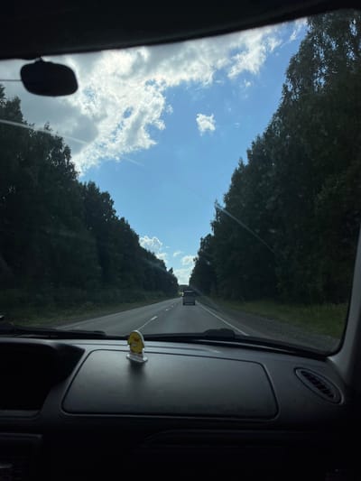The image shows a view from inside a vehicle traveling on a road bordered by dense trees. The sky above is blue with scattered white clouds. The dashboard of the car is visible in the foreground, with a small yellow figurine of a duck placed on it. The road ahead is paved and has lane markings. Another vehicle is visible in the distance on the same road. The overall scene suggests a journey through a forested area, possibly in a rural or suburban location. There are no discernible people or explicit interactions depicted. The image is taken during the daytime.