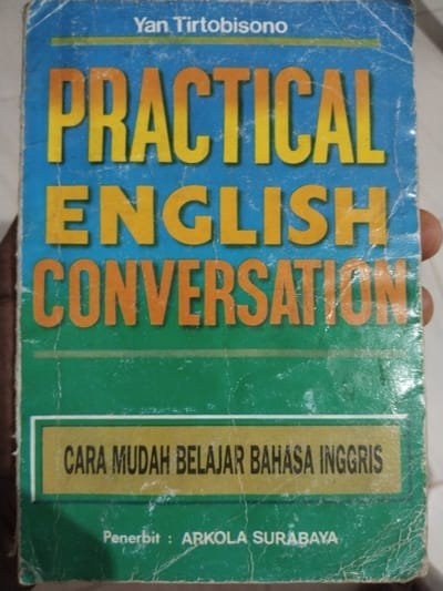 A book cover is shown, featuring the title "PRACTICAL ENGLISH CONVERSATION" in large, bold, yellow and orange letters against a blue background. Below this, in a green section of the cover, is a yellow banner with the text "CARA MUDAH BELAJAR BAHASA INGGRIS" in black lettering. At the bottom of the green section, the text "Penerbit : ARKOLA SURABAYA" is visible. The cover shows signs of wear, with some creasing and scuff marks, suggesting it is a well-used book. The lighting suggests it might be daytime, but there are no specific cues about the weather or time of day. The location context is Pekanbaru, Indonesia, but this is not visually evident from the image itself. The image is a close-up, focusing on the book's cover.
