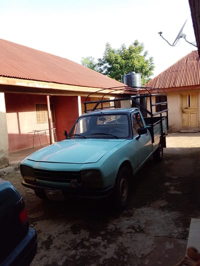 The image captures a light blue pickup truck parked in a courtyard. The truck, a Peugeot 504, has a black metal frame structure over its cargo bed, suggesting it's used for carrying goods. A large black water tank is visible on top of the structure, indicating water storage.
The setting appears to be a residential or commercial area in Mangu, Nigeria. Two buildings with corrugated red tin roofs are visible on either side of the courtyard. The building on the left has a red-painted wall with a window and some outdoor furniture. The building on the right has a cream-colored exterior and a yellow door.
The sky is bright and clear, suggesting it's daytime, likely morning or late afternoon due to the shadows cast. The ground is paved with a rough, textured surface that shows signs of wear and tear. A small part of another dark-colored vehicle is visible in the foreground on the left.
No people are visible in the image. The overall atmosphere is still and quiet, with no discernible activity taking place. The image is likely a general shot of the vehicle and its surroundings. There is no visible text in the image.