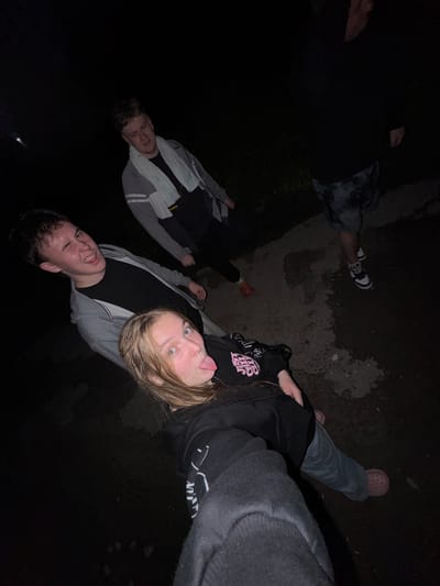 Four individuals are visible in the image, taken at night in what appears to be an outdoor, possibly urban or semi-urban environment with a wet, paved surface. The location is identified as Ryabinino, Russia.
In the foreground, a young person with blonde hair is sticking their tongue out, looking towards the camera. They are wearing a black hoodie with pink lettering and blue jeans. Next to them and slightly behind, a young person with short brown hair is winking and smiling, wearing a gray hooded jacket over a black t-shirt.
Behind these two, another young person stands, wearing a gray and white jacket with a light-colored towel draped around their neck. They appear to be looking down or away from the camera. Further back and to the right, a fourth individual is partially visible, wearing dark shorts with a camouflage pattern and black and white athletic shoes. Their lower body and feet are the most prominent features of this individual.
The ground is dark and appears to be wet in patches, suggesting recent rain or a damp environment. There is some grass or vegetation visible in the background to the left. The lighting is dim, indicative of nighttime, with some artificial light sources likely contributing to the illumination. The overall atmosphere is casual and informal.