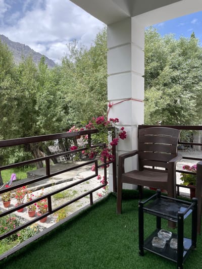 This image depicts an outdoor balcony area with a view of trees, mountains, and a clear sky. The balcony is furnished with a brown plastic chair and a small black side table that holds three glass ashtrays. The floor of the balcony is covered with artificial green turf. A railing separates the balcony from a lower area where several potted plants are visible. In the background, a mountain range can be seen through a dense canopy of green trees. The architecture of the building includes a white pillar with decorative molding. The scene appears to be a tranquil setting, possibly a guesthouse or hotel balcony in a mountainous region.