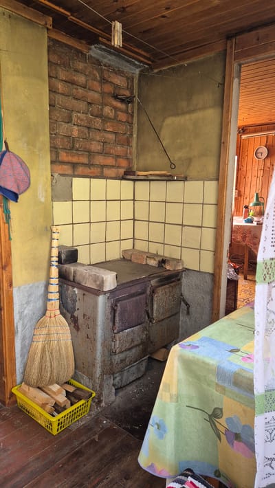 The image depicts a rustic interior scene, likely in a rural or older dwelling, possibly in the village of Mashezero, Russia. The foreground features a worn wooden floor. To the left, a broom made of straw stands upright in a bright yellow plastic basket filled with cut firewood. Next to the broom, a portion of a red and blue patterned hat hangs from a wooden doorframe.
Dominating the center of the image is an old, metal stove with a tiled backsplash. The stove appears to be made of cast iron with visible doors and a flat top. Several bricks are placed on top of the stove. The tiled area behind the stove is a pale yellow color. Above the tiled area, a brick wall is visible, with a thin black metal rod extending from the wall towards the right.
To the right of the stove, a table is partially visible, covered with a tablecloth featuring a colorful floral pattern in shades of green, yellow, blue, and pink. A white lace curtain hangs on the right edge of the image. Beyond this, an open doorway reveals another room with wooden walls and ceiling, and a table with a green lamp. The overall impression is one of a functional, lived-in space.