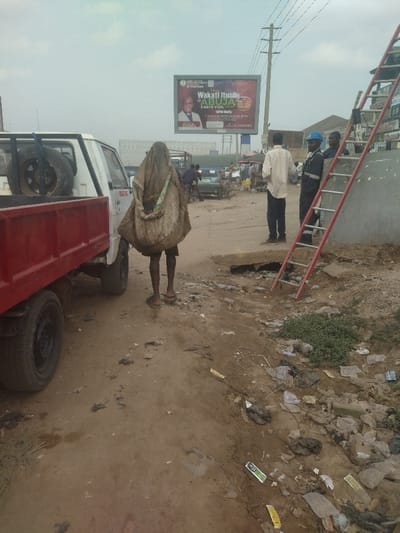 The image shows a scene on a dirt road in Way, Nigeria, located at 9.029 latitude and 7.584 longitude. A white truck with a red bed is parked on the right side of the image. There are three people walking in the scene. The person in the center is wearing a long, loose-fitting, beige garment covering their entire body except for their hands and feet. The person on the left is dressed in a white t-shirt and black trousers. The person on the right is dressed in blue and brown clothes, with a red hard hat on their head. There is a ladder leaning against the wall of a building on the right side of the scene. The ground is covered in trash and debris. There is a billboard in the background, with a message in a foreign language. The scene is an unkempt urban area.