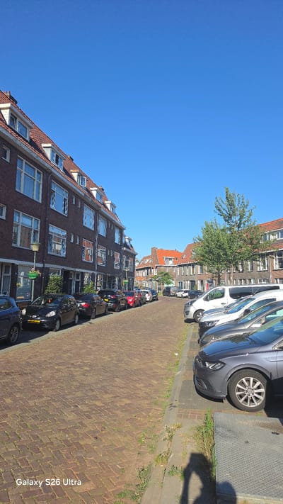This media file depicts a street scene in Vlaardingen, Netherlands. The image is oriented vertically, showing a clear blue sky with a row of brick buildings lining a cobblestone street. Several cars are parked along the street, indicating a residential or urban area. The sunlight suggests it is daytime, likely morning or afternoon, given the shadows. The buildings appear to be apartments or townhouses, with multiple windows and red-tiled roofs, some with skylights. A tree is visible between two buildings, adding a touch of greenery to the urban landscape. The cobblestone street is laid in a herringbone pattern. The overall impression is one of a quiet, sunny day in a European city. The text "Galaxy S26 Ultra" is visible in the lower right corner, which is likely a watermark from the device used to capture the image. There are no people visible in the image.