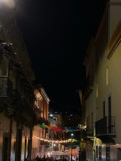This image depicts a narrow, bustling pedestrian street at night in Guanajuato City, Mexico. The street is flanked by multi-story colonial-style buildings; the left structure features an aged, textured facade with several ornate wrought-iron balconies, while the right building has a smoother, lighter-toned facade with simpler balconies. The dark night sky is visible above, with distant city lights illuminating hills in the background.

The street is extensively illuminated by multiple strings of warm-toned bulb lights suspended across its width, supplemented by street lamps, one clearly visible on the right. Multi-colored 'papel picado' banners, in shades of red, orange, yellow, green, and blue, are strung above the string lights. A dense crowd of people occupies the lower section of the street, indicating active pedestrian traffic. Within the crowd, several large, light-colored patio umbrellas or canopies are visible, suggesting outdoor dining establishments or vendor stalls. A white street sign with blue text, partially reading "SALAO," and a yellow pedestrian crossing sign are visible on the right side of the street. Electrical or communication wires traverse the sky between buildings.