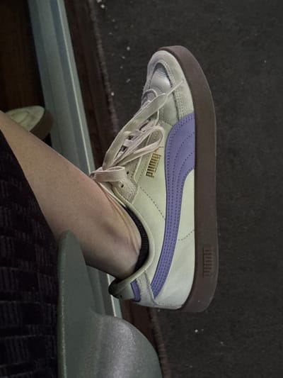 A close-up photograph shows a person's leg wearing a white Puma sneaker with a purple stripe. The sneaker has white laces, a brown sole, and the brand name "PUMA" in gold on the side. The leg is resting near a green hat. The background includes a dark floor and a light-colored metal or plastic surface. The location is not discernible from the image.