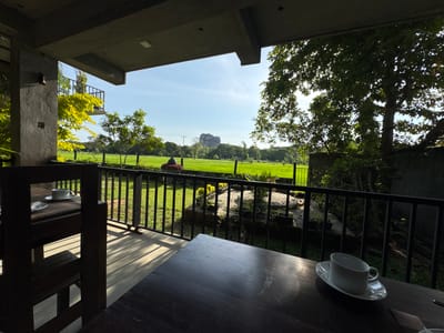 A landscape shot taken from an outdoor patio in Sigiriya, Sri Lanka. A black, metal-railed fence runs along the edge of the patio, which overlooks a green field and trees. In the distance, a large rock formation can be seen. The patio itself is covered by a gray ceiling.
There are two tables on the patio. The table in the foreground is made of dark wood and has a white cup and saucer sitting on it. The table in the background is also made of dark wood and has a white cup and saucer on it. A wooden chair is positioned next to the table in the background.