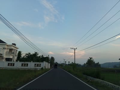A black truck drives down an asphalt road lined with white markings on the left side, towards a landscape dominated by green fields and palm trees under a pale blue sky with thin clouds. On the left side of the road, a modern white house with a black roof and multiple stories is visible behind a white fence with rectangular openings. Several utility poles with electrical wires are present along the road, and in the distance, a gently sloping hillside can be seen. The scene appears to be a rural or semi-rural area in Uleejalan, Indonesia, possibly at dusk or dawn due to the soft lighting.